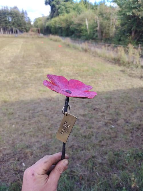 A hand holding a metal poppy outdoors. The flower head is bright red with a dark center, attached to a black stem with a brass-colored hanging tag engraved.