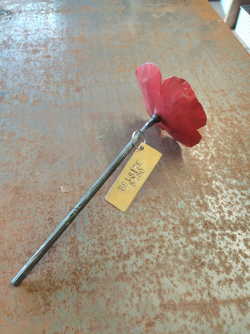 A handmade metal poppy flower with a textured red bloom and a long silver stem, resting on a rustic, rusted metal surface. A small golden tag is attached to the stem.