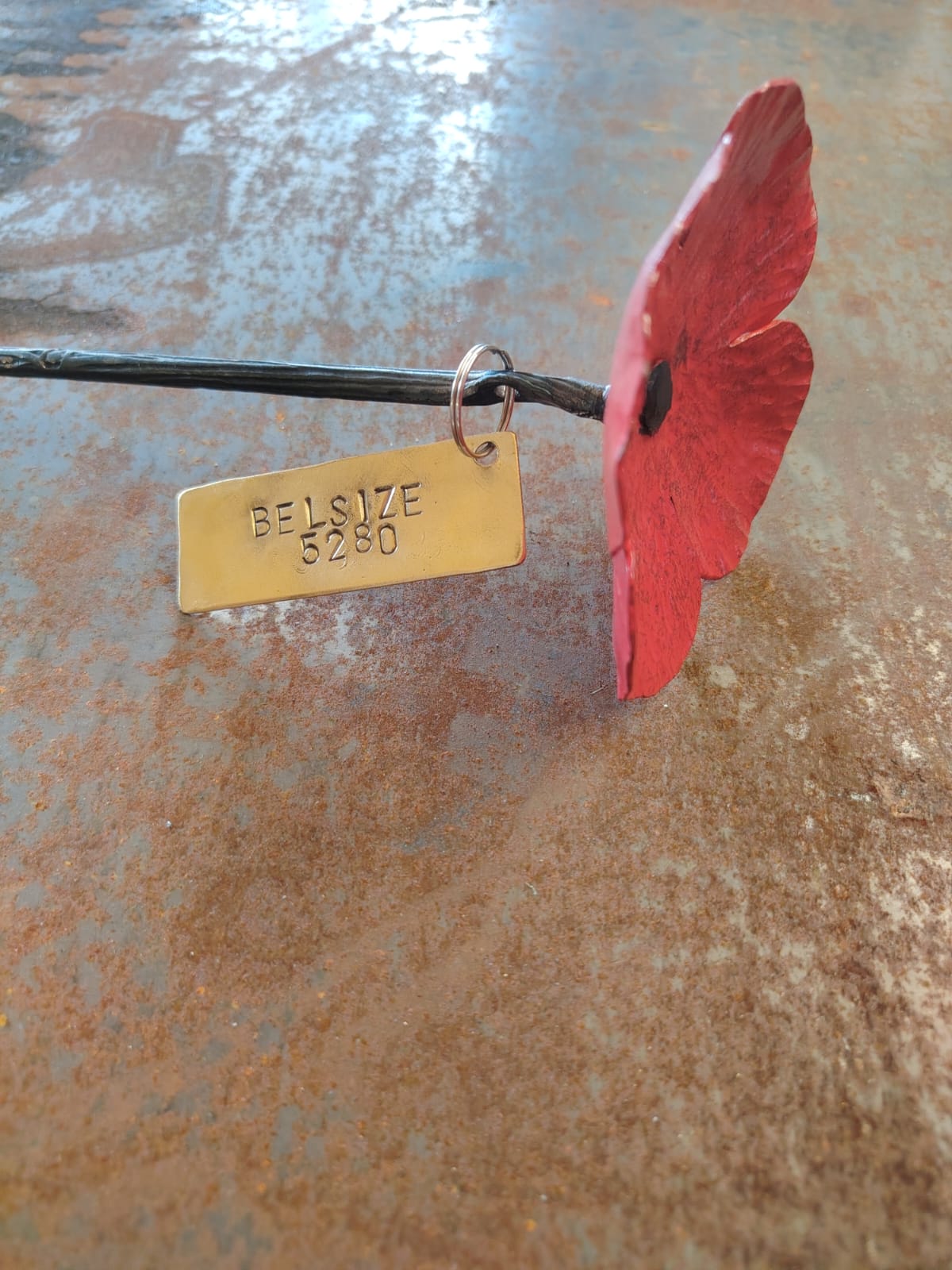 A handmade metal poppy remembrance flower with a stamped brass size tag reading 'BELSIZE 5280', set against a rustic, textured rusty brown background.